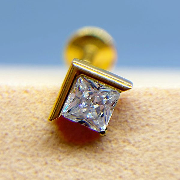 Close-up of a gold stud earring with a square clear gemstone against a blurred blue and beige background