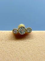 Three gold studs with clear gemstones on skin against a blue background