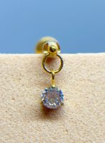 Gold stud earring with a dangling clear gemstone on beige fabric against a blue background