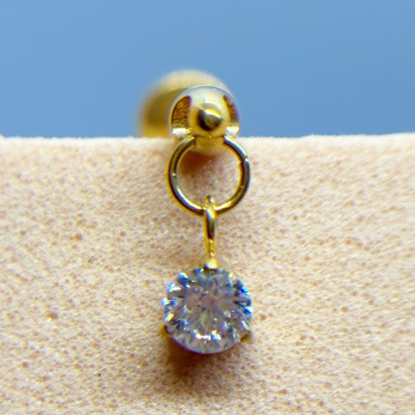Gold stud earring with a dangling clear gemstone on beige fabric against a blue background
