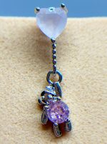 Close-up of a belly button piercing with a heart-shaped gem and dangling purple turtle charm