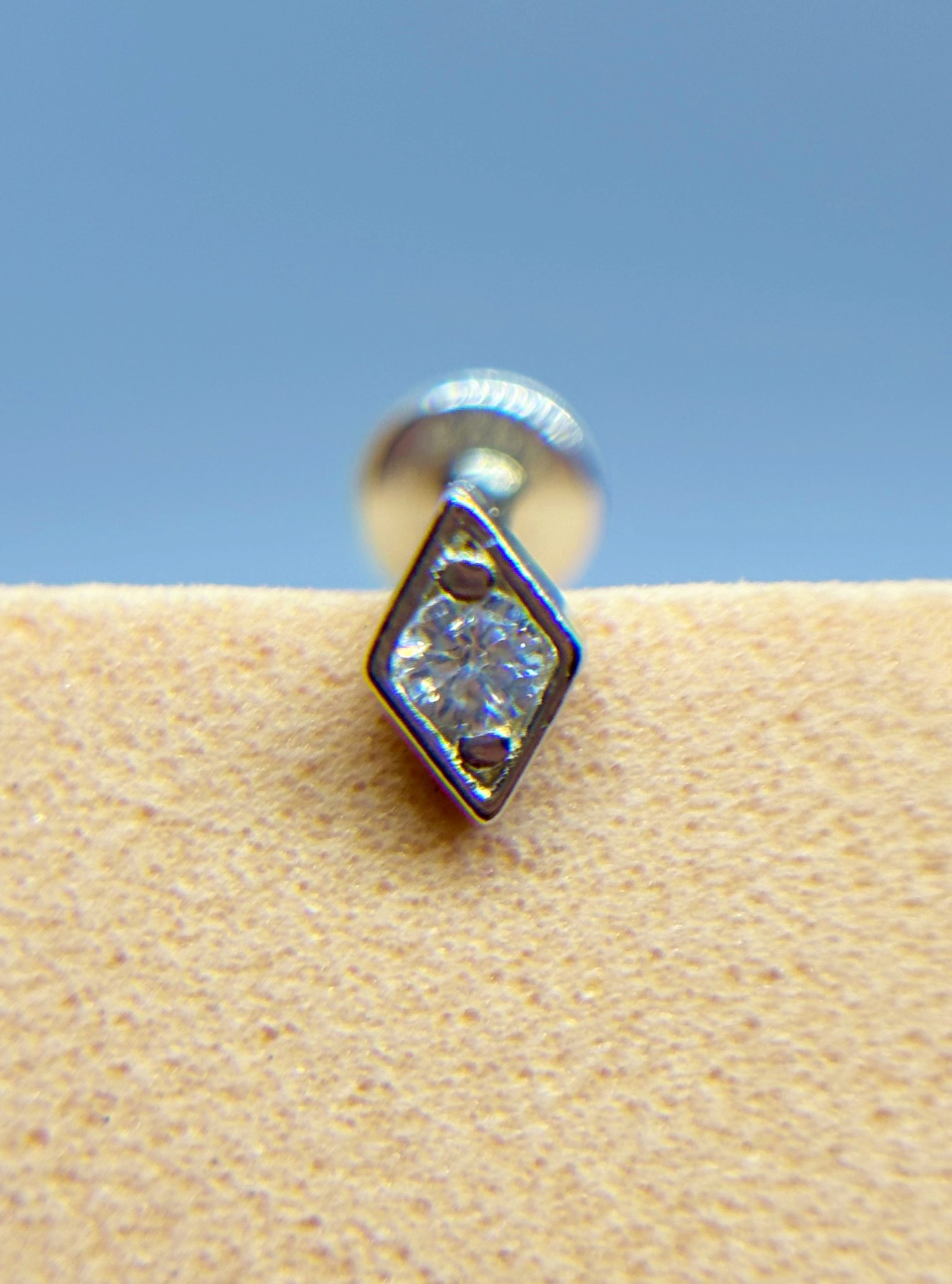 IMG_9440 Close-up of a diamond-shaped stud earring with a clear stone against a blue background