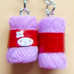 Two tiny purple-and-red knitted-looking earrings with metal hooks, one showing a small white face design